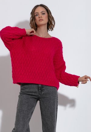 Puchaty, oversize sweter w jaskrawym różu, z luźnym wzorem splotu, szerokim dekoltem i długimi rękawami. Zestawiony z szarymi dżinsami z wysokim stanem.