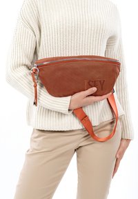 Bruine leren heuptasje met een gestructureerd oppervlak, zilveren ritssluiting en verstelbare oranje band, vastgehouden door een persoon die een witte geribbelde trui draagt.