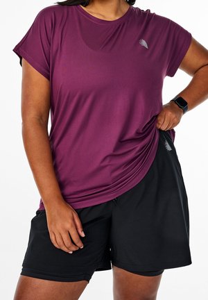 Osoba v tmavě červeném atletickém tričku s krátkými rukávy a černých šortkách, s chytrými hodinkami na levém zápěstí, si upravuje tričko.