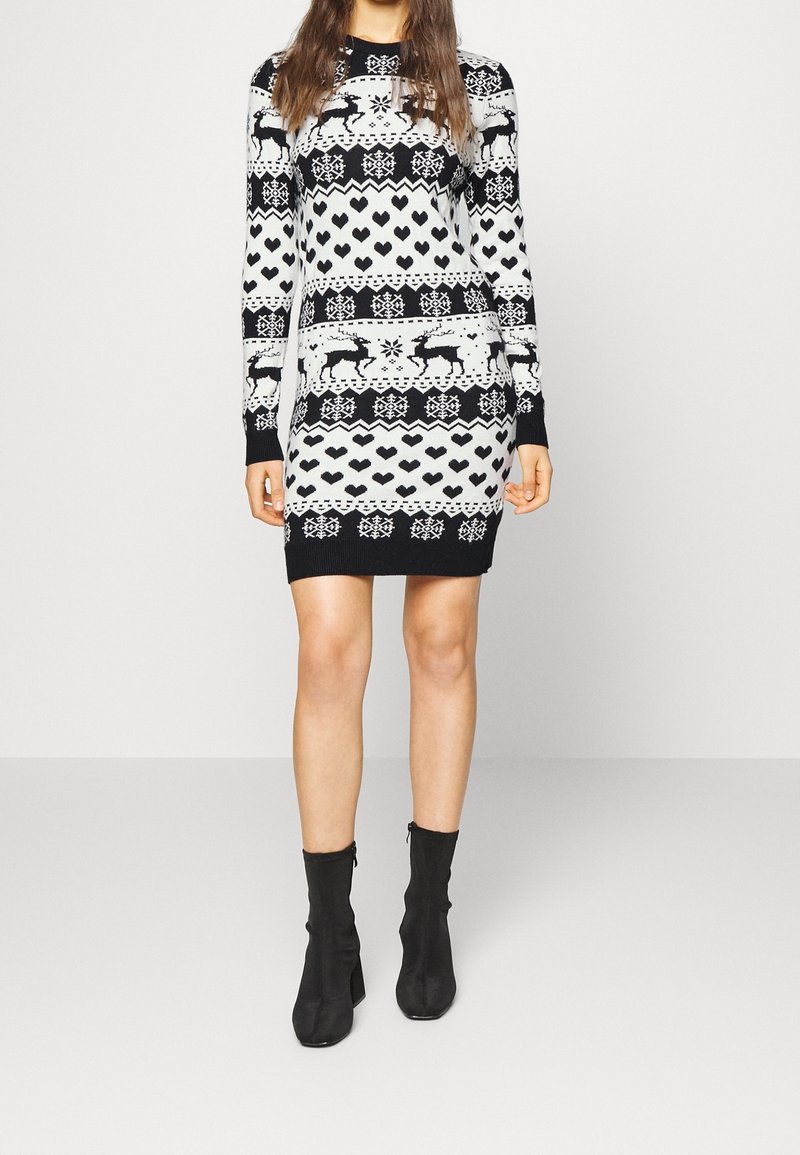 Femme portant une robe pull noire et blanche tricotée avec des motifs de rennes, de cœurs et de flocons de neige, associée à des bottines noires.