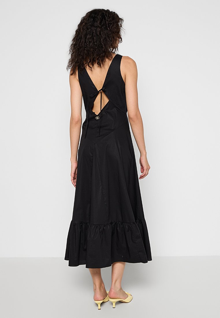Czarna sukienka bez rękawów z niskim dekoltem, zdobiona wiązaniem i falbaniastym dołem. Gładki materiał o matowym wykończeniu.
