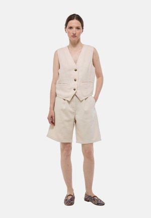 Gilet en lin beige avec un décolleté en V profond et trois boutons, associé à un short beige ample arrivant aux genoux. Porté avec des chaussures à enfiler à motifs.