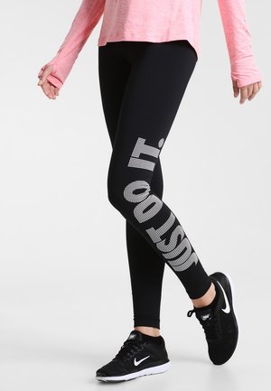 Leggings noirs avec une finition texturée portant l'inscription "JUST DO IT" en blanc sur la jambe gauche. Associés à des chaussures de sport noires et un haut rose.