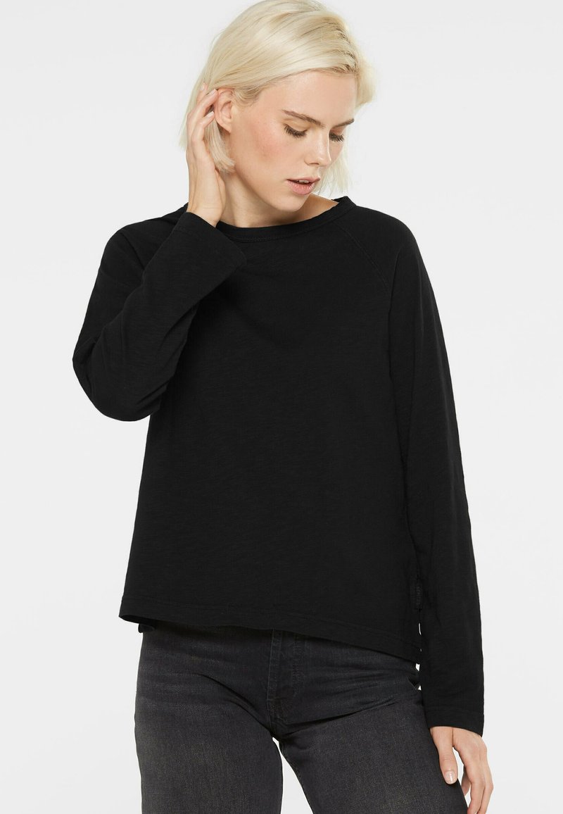 Blonde Frau, die ein schwarzes Langarmshirt und dunkle Jeans trägt, mit einer Hand ihr Haar berührt und nach unten schaut.