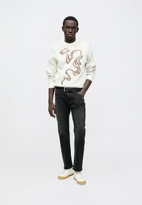 Light cream sweater with a wavy brown design, paired with dark jeans and white sneakers with a beige sole. Simple, form-fitting design.