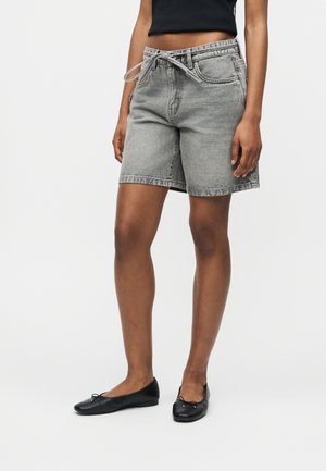 Femeie purtând pantaloni scurți din denim gri decolorați cu o curea legată, top negru fără mâneci și balerini negri, stând pe un fundal alb simplu.