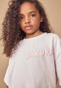 Šviesiai pilka medvilninė marškinėlių su švelniai rožine ir žalia siuvinėta užrašu "SAKURA". Trumpomis rankovėmis, laisvo kirpimo, su minkštu tekstūru.