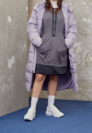 Osoba w jasnofioletowym długim puchowym płaszczu na szarej sukience z czarnym wykończeniem, białych skarpetkach i białych trampkach, stojąca na niebieskim dywanie.