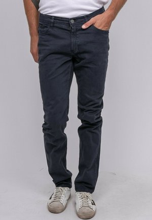 Homme portant un jean bleu foncé ajusté et des baskets blanches avec des semelles usées, debout avec une main dans la poche devant un fond uni.