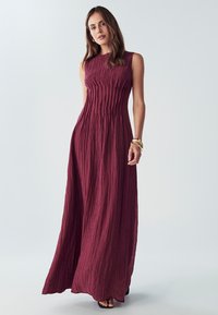 Bordová maxi šaty bez rukávov s textúrovaným, plisovaným dizajnom a rovnou siluetou. Nosené s čiernymi sandálmi a minimálnymi doplnkami.