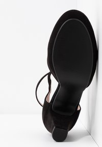 Chaussure à talon haut en daim noir avec un bout pointu, un dos ouvert et une sangle à la cheville, présentant une semelle en caoutchouc texturée.