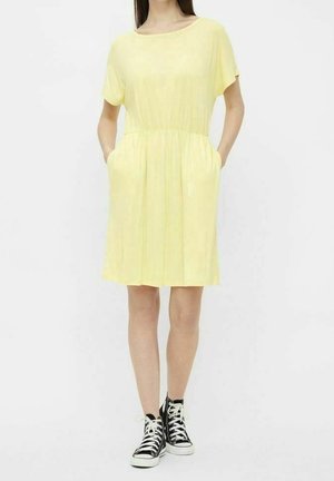 Femme portant une robe jaune à manches courtes, longueur genou, avec des poches, et des baskets montantes noires, debout devant un fond uni.