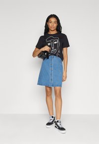 Zwarte T-shirt met grafische afbeelding van een gezicht in het wit, gecombineerd met een lichtblauwe denimrok met knopen aan de voorkant. Zwarte sneakers maken de outfit compleet.
