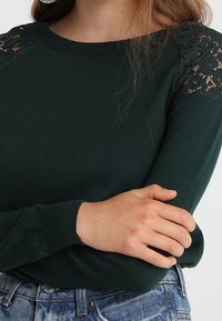 Zelený pletený svetr s krajkovými akcenty na ramenou, kulatým výstřihem a dlouhými rukávy, v kombinaci s světle modrými džínami.