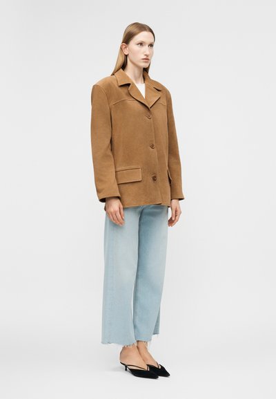 Γυναίκα που φοράει ένα καφέ παλτό με κουμπιά, ανοιχτό μπλε τζιν παντελόνι με φαρδιά μπατζάκια και ξεφτισμένα τελειώματα, καθώς και μαύρα παπούτσια με χαμηλό τακούνι και λουράκι στο πίσω μέρος, στέκεται μπροστά σε ένα λευκό φόντο.