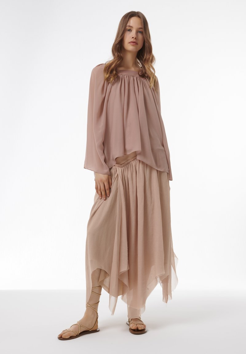 Eine blushfarbene, fließende Chiffonbluse mit gerafftem Ausschnitt und luftigen Ärmeln, kombiniert mit einem passenden High-Low-Rock und Riemchensandalen.