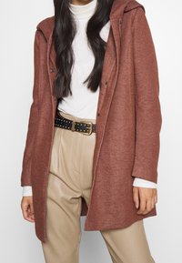 Abrigo marrón texturado con capucha, con botones y un corte relajado. Lleva puesto un jersey de cuello alto blanco y se complementa con pantalones beige.