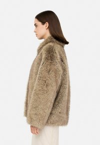 Giacca in pelliccia sintetica color marrone chiaro, di taglio oversize con un ampio colletto. Texture morbida e silhouette ampia, vista di lato, indossata sopra un vestito di tessuto leggero.