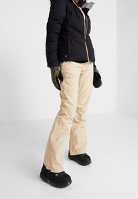 Veste matelassée noire avec une fermeture éclair et une capuche, pantalon beige clair, gants verts et bottes imperméables noires avec des accents verts.