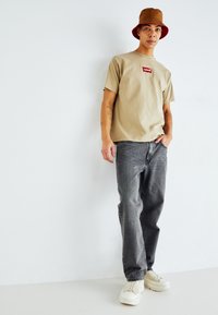 Beige bomulds T-shirt med et rødt Levi's-logo, parret med grå denimjeans og hvide sneakers. Modellen bærer en brun bucket hat.
