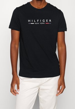 Mężczyzna w czarnej koszulce z krótkim rękawem z napisem "HILFIGER NEW YORK" i logo, ubrany w białe spodnie, stoi na jednolitym tle.