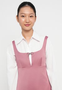 Roze jurk met een vierkante halslijn en diepe uitsnijding, met een zilveren strikaccent in het midden. Gelaagd over een witte blouse met kraag.