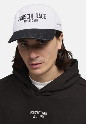 Witte baseballpet met "PORSCHE RACE AMERICANA" in het zwart geborduurd, met een contrasterende donkere klep, gedragen met een zwarte hoodie met witte tekst.