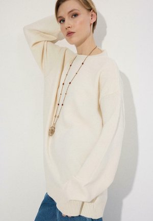 Jeune femme aux cheveux blonds courts, portant un pull oversize crème, un long collier de perles et un jean bleu, posant sur un fond clair.