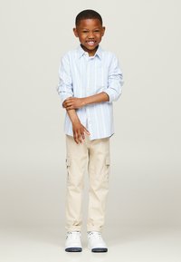 Camicia azzurra a righe con bottoni, pantaloni cargo beige con tasche laterali e sneakers bianche. La camicia ha una tasca sul petto e le maniche arrotolate.