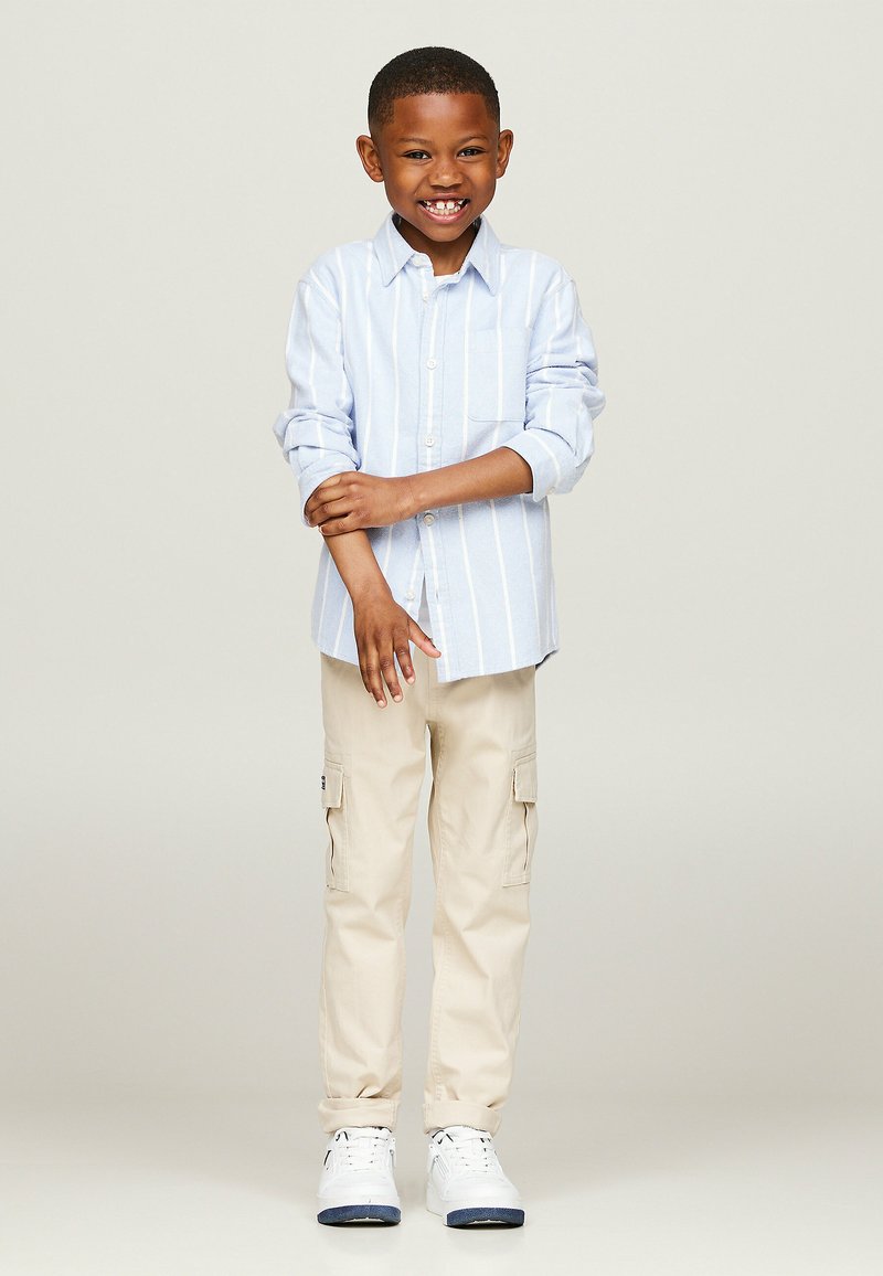 Camicia azzurra a righe con bottoni, pantaloni cargo beige con tasche laterali e sneakers bianche. La camicia ha una tasca sul petto e le maniche arrotolate.