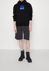 Sudadera negra con logo azul, pantalones cortos grises y botas de combate negras. El material parece suave; la sudadera cuenta con un bolsillo canguro y cordón ajustable.