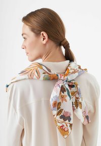 Zijden sjaal met een bloemmotief in oranje, blauw en groen, om de nek geknoopt. G gedragen over een lichtkleurige blouse.