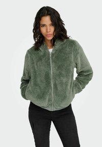 Zielona, puszysta kurtka bomber z długimi rękawami i zamkiem błyskawicznym, o miękkiej teksturze i elastycznych mankietach, zestawiona z czarnymi spodniami.