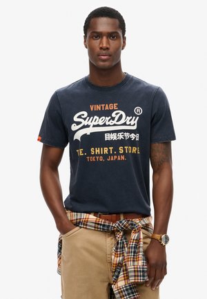 Marineblå bomulds T-shirt med hvid og orange "SuperDry" logo tekst, rund halsudskæring, korte ærmer, båret med beige bukser og en ternet skjorte bundet om taljen.