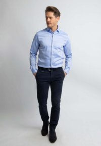 Lichtblauwe overhemdbloes met een soepele textuur, gecombineerd met donkerblauwe broek. De outfit heeft een zwarte riem en donkere schoenen.