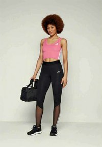 Fit Frau mit Afro, die ein pinkes Crop-Top, schwarze Leggings mit Mesh-Einsätzen und schwarze Sneakers trägt, während sie eine schwarze Sporttasche hält, und vor einem schlichten Hintergrund steht.