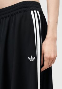 Schwarze Sporthose mit weißen drei Streifen und Adidas-Logo an der Seite, zeigt einen Teil der Taille und eine Hand einer Person.