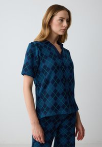 Top blu navy a maniche corte con motivi a diamante verde e teal. Presenta un collo a V e una vestibilità rilassata. Tessuto morbido con un design di colori complementari.