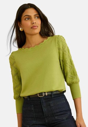 Žena s dlhými tmavými vlasmi, ktorá nosí zlaté náušnice, zelený sveter s textúrou a vlnkovým výstrihom a tmavé rifle s vysokým pásom a čiernym opaskom.