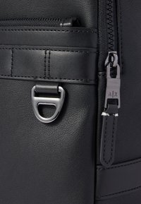 Gros plan sur un sac à dos en cuir noir, mettant en avant une fermeture éclair argentée, un détail en métal en forme de D, et des coutures contrastantes sur la surface.