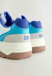 Sneakers sportive con una toma blu e teal, dettagli in suede grigio, suola intermedia bianca e una suola esterna marrone texturizzata con un motivo a freccia distintivo.