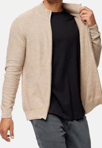 Hombre vistiendo un suéter cremoso con cremallera sobre una camiseta negra y pantalones gris oscuro, con una mano tirando del suéter hacia afuera.