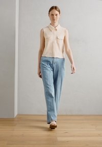 Canotta beige chiara senza maniche con due tasche frontali, abbinata a jeans a gamba larga blu chiaro e sandali marroni, su uno sfondo neutro.