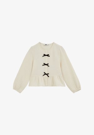 Blouse crème à manches longues avec encolure ronde, trois nœuds noirs sur le devant et ourlet bas à volant froncé.