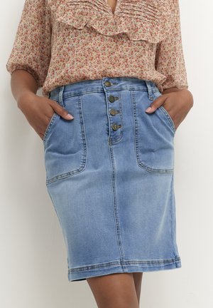 Frau trägt einen hellblauen Jeansrock mit vier vorderen Knöpfen und großen Taschen, kombiniert mit einer geblümten Bluse mit Rüschen und 3/4-Ärmeln.