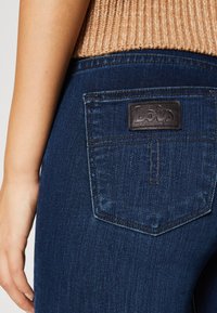 Tmavě modré džíny vyrobené z denimu s koženou nášivkou embosovanou nápisem "Lais," a zadní kapsou zvýrazněnou kontrastním prošíváním. Viditelná strukturovaná tkanina.