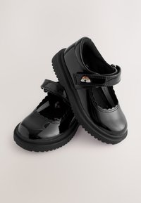 Paar glänzende schwarze Mary Jane Schuhe mit dicken Sohlen und einem Regenbogen-Aufnäher am Riemen, auf hellem Hintergrund platziert.