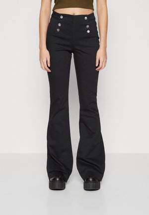 Persona con pantalones negros acampanados de talle alto con botones plateados decorativos y zapatos de plataforma negros, con top corto color oliva visible.