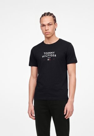 Jeune homme aux cheveux tressés portant un t-shirt noir Tommy Hilfiger et un pantalon noir, debout sur un fond blanc uni.