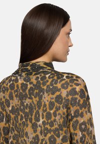 Przezroczysta bluzka z nadrukiem w cętki w złotych i czarnych odcieniach, z wysokim kołnierzem i teksturowaną tkaniną. Detale widoczne przy dekolcie.